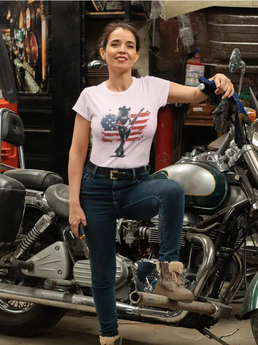 Confident woman in a garage wearing a patriotic T-shirt with silhouette of a dancing cowgirl in front of a U.S. flag – countrywear by Rugged Rhythm.